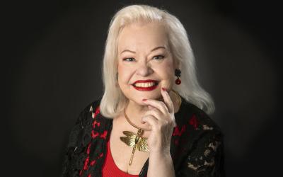 Anneli Saaristo. Vaaleatukkainen nainen jolla on punaista ja mustaa vaatetta, sekä kaulassa kultaista sudenkonteroa esittävä kaulakoru.