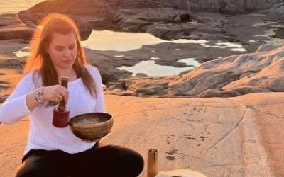 Nainen istuu rantakalliolla käsissään astioita, takana auringonlasku