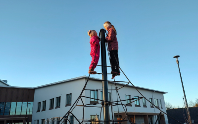 Kaksi lasta seisoo rakennuksen edessä olevassa lasten kiipeilytelineen huipulla. Taustalla vaaleahko koulurakennus ja sininen taivas.