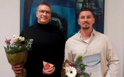 kaksi palkittua miestä seisoo kukkien ja palkinnon kera taulun edessä