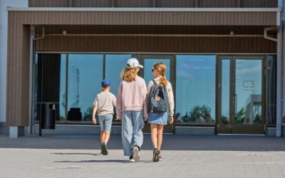 Kolme lasta kävelee kohti koulurakennusta aurinkoisena päivänä.