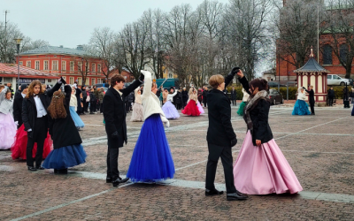 Nuoria tanssii näyttävissä juhlamekoissa, takeissa ja frakeissa ulkona torilla.