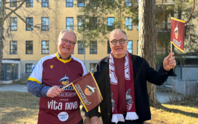 Kaupunginjohtaja Atso Vainio ja elinkeinojohtaja Jarkko Heinonen hymyilevät ja heiluttavat Korihaiden viininpunaisia viirejä ulkona otetussa kuvassa. Taustalla keltainen virastotalo.