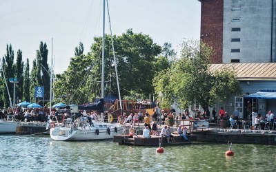 Kuvassa kaupunginlahti, veneitä, pakkahuoneen ranta ja ihmisiä kahvilassa