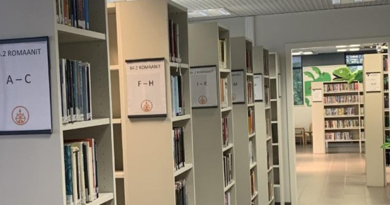 Kuva kirjaston sisäpuolelta. Kuvan vasemmalla reunalla on kirjahyllyjä, joiden kylteissä on kirjainopasteet.
