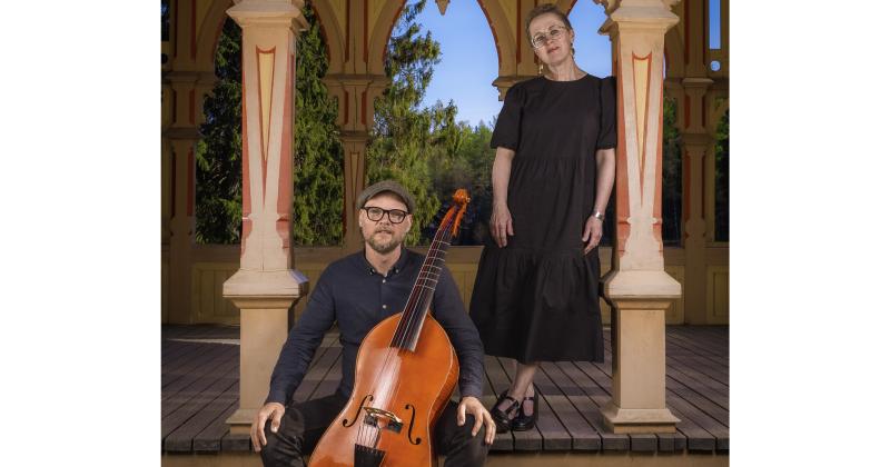 Mikko Perkola ja Annamaria Hannonen ovat vanhan puurakennuksen kuistilla. Perkolan sylissä on jousisoitin viola de gamba.
