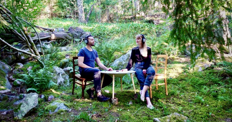 Kaksi ihmistä istuvat tuoleilla metsässä pyöreän pöydän äärellä silmät suljettuina. Molemmilla on kuulokkeet päässä.