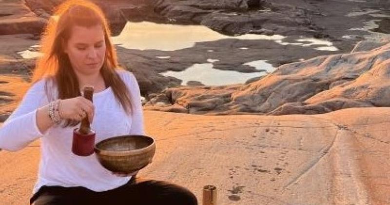 Nainen istuu rantakalliolla käsissään astioita, takana auringonlasku