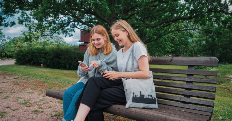 Kaksi teini-ikäistä tyttöä istuvat vierekkäin ruskealla puiston penkillä ja molemmat katsovat hymyillen puhelimiaan. Taustalla näkyy kesäisen vihreää puistomaisemaa.