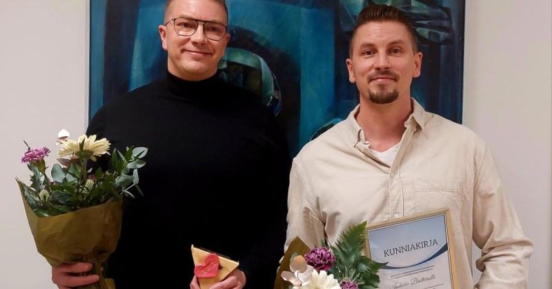 kaksi palkittua miestä seisoo kukkien ja palkinnon kera taulun edessä