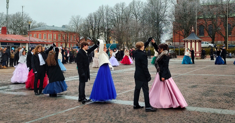 Nuoria tanssii näyttävissä juhlamekoissa, takeissa ja frakeissa ulkona torilla.