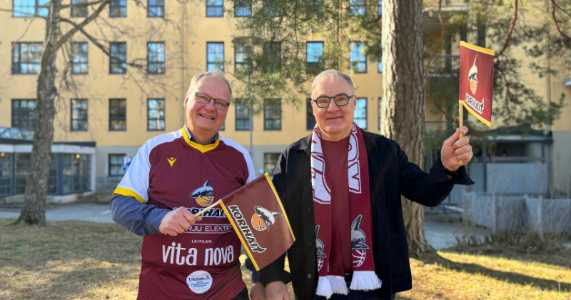 Kaupunginjohtaja Atso Vainio ja elinkeinojohtaja Jarkko Heinonen hymyilevät ja heiluttavat Korihaiden viininpunaisia viirejä ulkona otetussa kuvassa. Taustalla keltainen virastotalo.