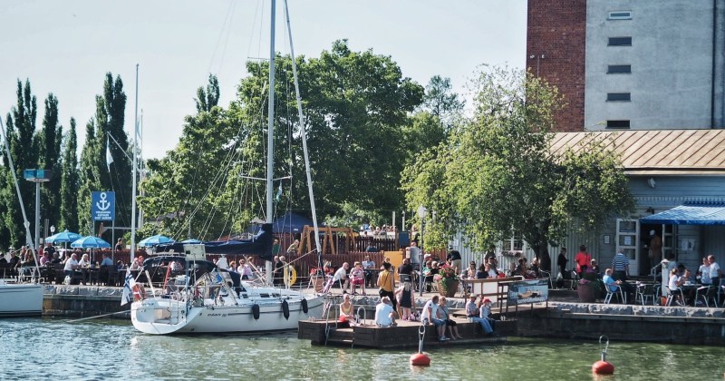 Kuvassa kaupunginlahti, veneitä, pakkahuoneen ranta ja ihmisiä kahvilassa