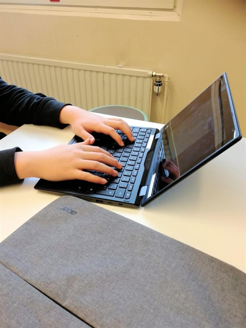 Oppilaan kädet Chromebookin näppäimistöllä, jonka vieressä on suojapussi.